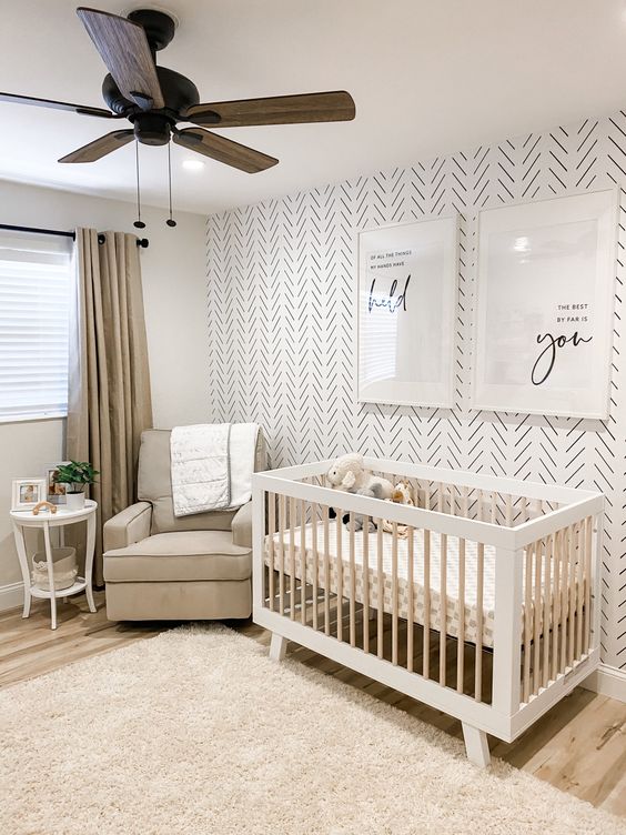 a chic modern nursery with a print accent wall, a grey chair and a white crib, a neutral rug, grey curtains and a wooden gfan plus some prints