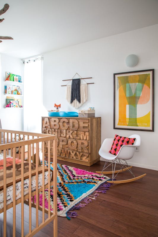 a colorful boho nursery with a stained crib and a dresser with letters carved, a white rocker, a colorful rug, bedding, colorful books on the ledges