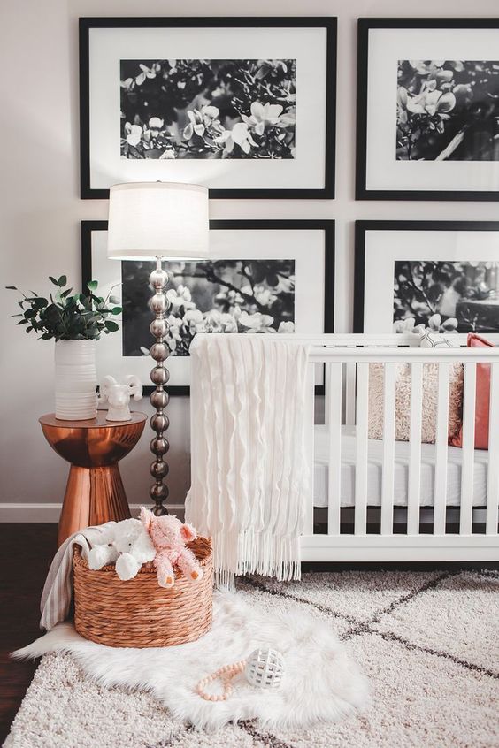 a contemporary nursery with a black and white gallery wall, a white crib, layered rugs, a basket with toys, a copper table and a floor lamp