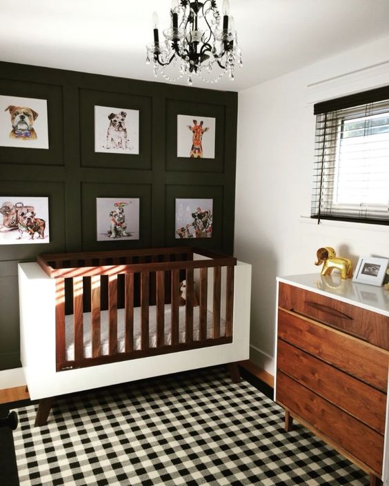 a cool gender neutral nursery with a black accent wall with prints, a plaid rug, a white and stained crib and a matching dresser plus a chic chandelier