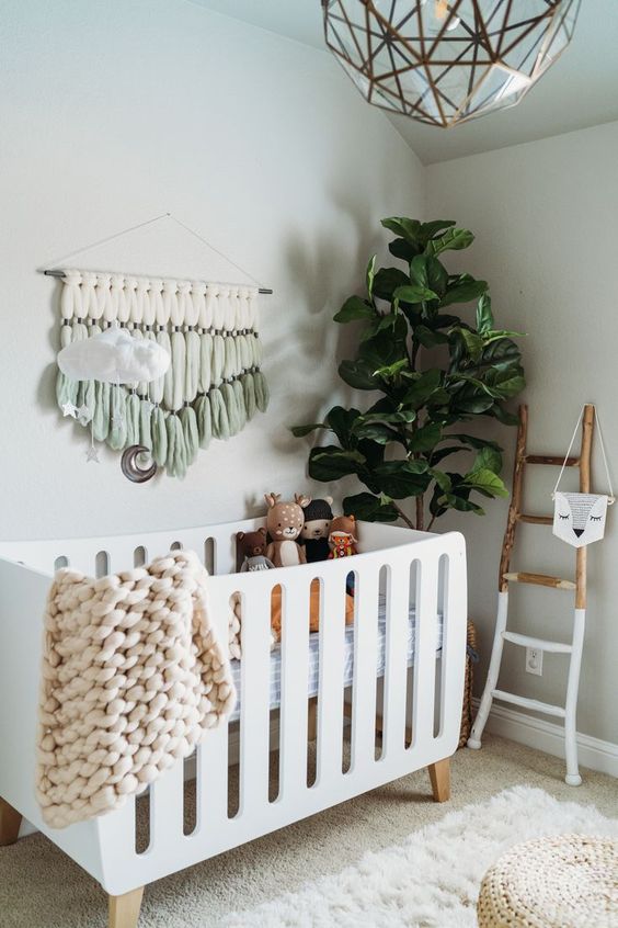 a cozy boho nrusery with a white crib, a painted ladder, some lovely boho decor, toys, poufs and a statement plant is very welcoming