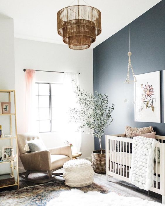 a delicate and cool contemporary nursery with a black accent wall, a white crib, a tan rocker chair, an open shelving unit, a fringe chandelier and a tree