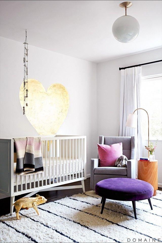 a gorgeous modern nursery with a white crib, a gold heart on the wall, a grey chair with a pink pillow and a purple ottoman is cool