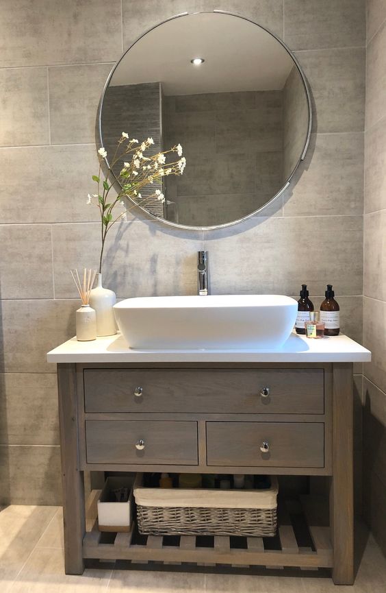 a grey tile bathroom with a taupe vanity, a white sink, a round mirror, baskets for storage is a very chic and cool idea to rock