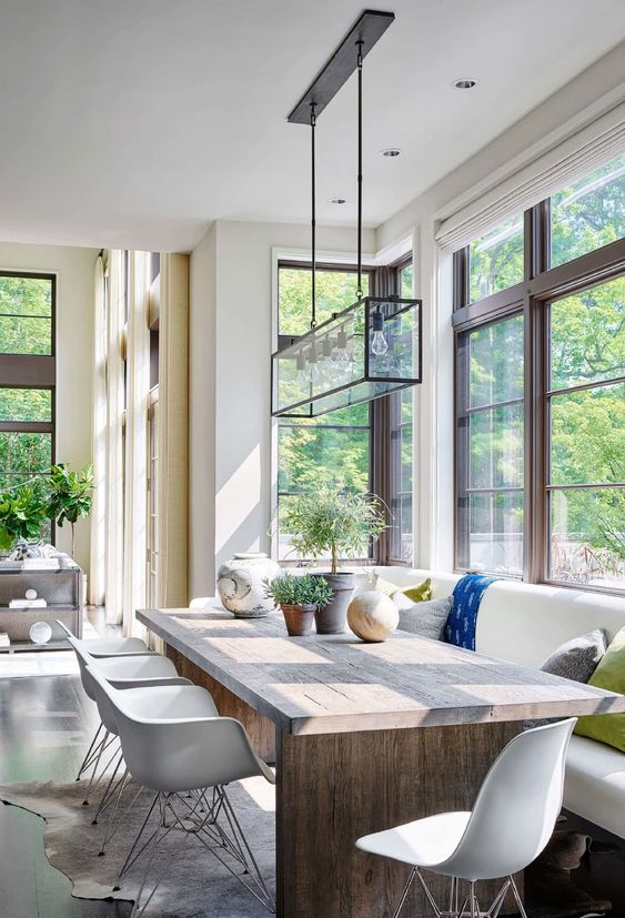 a light-filled contemporary dining room with forest views, a built-in bench, a wooden table, white chairs, a glass chandelier and potted plants