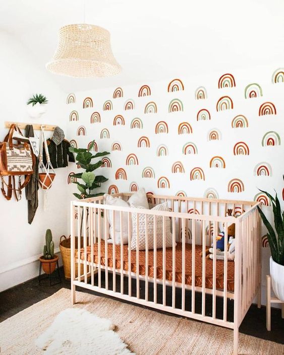 a lovely boho nursery with a rainbow accent wall, a pink crib, bright bedding, layered rugs, a bead pendant lamp and statement plants