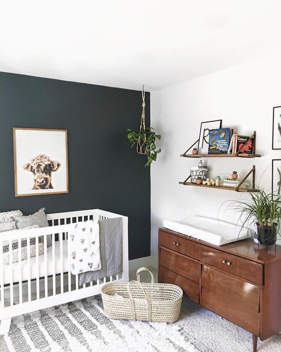 a lovely modern nursery with a black accent wall, a white crib, a rich-stained dresser, open shelves, potted plants, a boho rug and printed bedding