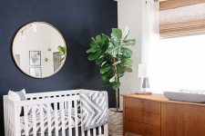 a mid-century modern nursery with a navy accent wall, a crib with printed bedding, a rich-stained changing table, a colorful printed rug and a chic chandelier