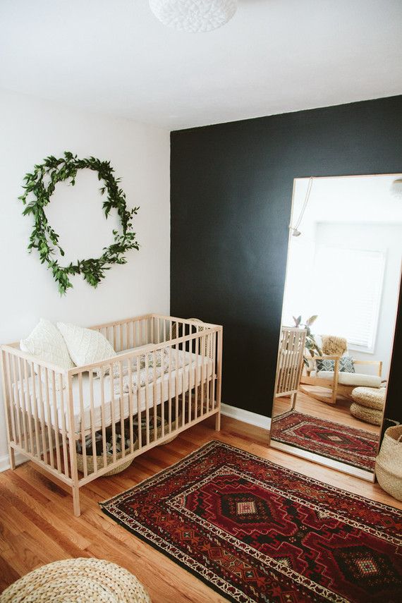 a modern boho nursery with a black accent wall, a stained crib and chair, a floor mirror, a printed rug, neutral bedding and a greenery wreath