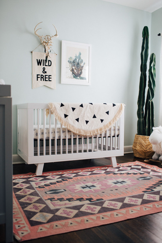 a modern boho nursery with a printed rug and blanket, a cool desert gallery wall, a statement cactus in a wooden planter and a grey dresser