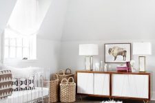 a modern glam nursery with an acrylic crib, a fluffy pouf, a white and stained credenza, baskets for storage and a purple statement rug
