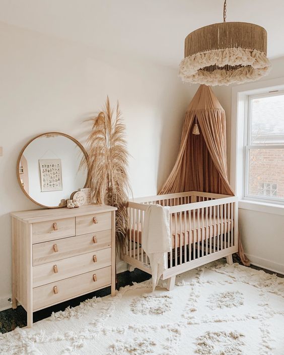A neutral boho nursery with stained furniture, a rust colored canopy, pampas grass, a round mirror and a cool tassel pendant lamp