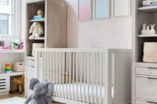 a pretty contemporary nursery with a pink printed wall, neutral furniture, layered rugs, lovely toys and a desk for the kid