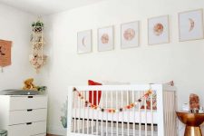 a pretty modern nursery with white furniture, moon prints, a lamp and a fan, a hanging shelf with toys, layered rugs and a shiny side table