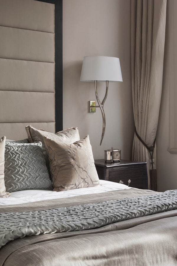 a refined taupe bedroom with a bed with an oversized upholstered headboard, a dark nightstand, a chic sconce and taupe and white bedding