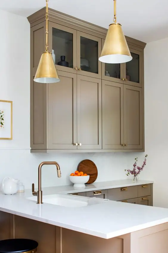 A refined taupe kitchen with shaker cabinets   mostly lower and a couple of upper ones, white stone countertops and a backsplash, gold pendant lamps