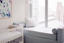 a simple contemporary nursery with a white crib, a colorful artwork, a grey daybed, a neutral rug and a view of the city