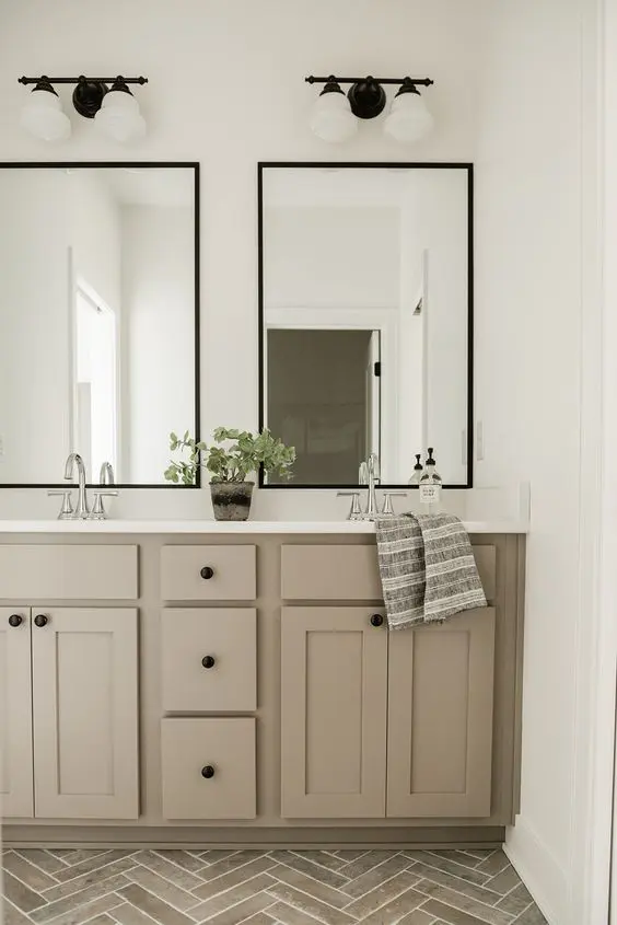 a stylish bathroom with a grey chevron tile floor, a taupe double vanity, two mirrors, sconces and a potted plant is a very chic idea