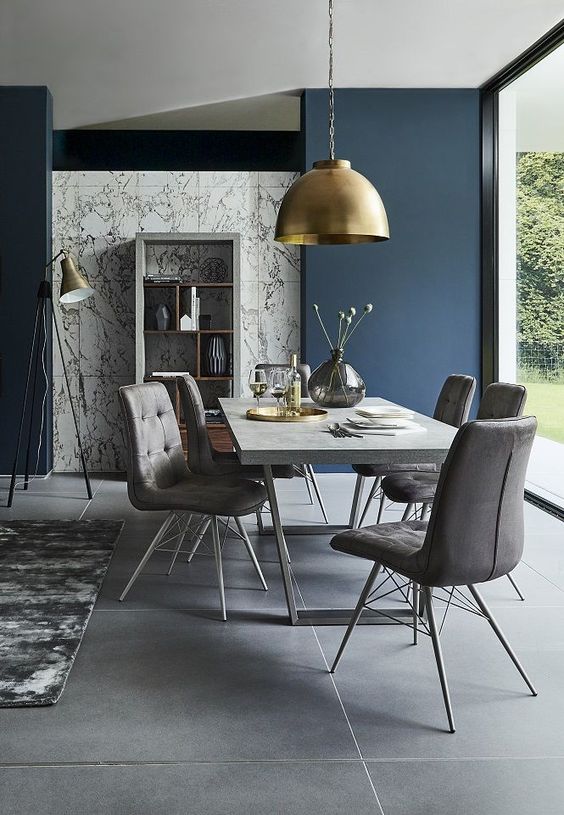 a stylish contemporary dining room with a glazed wall, a concrete table, grey upholstered chairs, a gold pendant lamp and candles