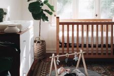 a stylish modern boho nursery with a stained crib, a stained and white dresser, a printed rug, a house-shaped shelf and statement plants
