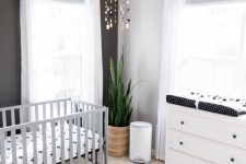 a stylish monochromatic nursery with a white dresser, a grey crib, a black accent wall, a printed rug and bedding, a mobile and white curtains