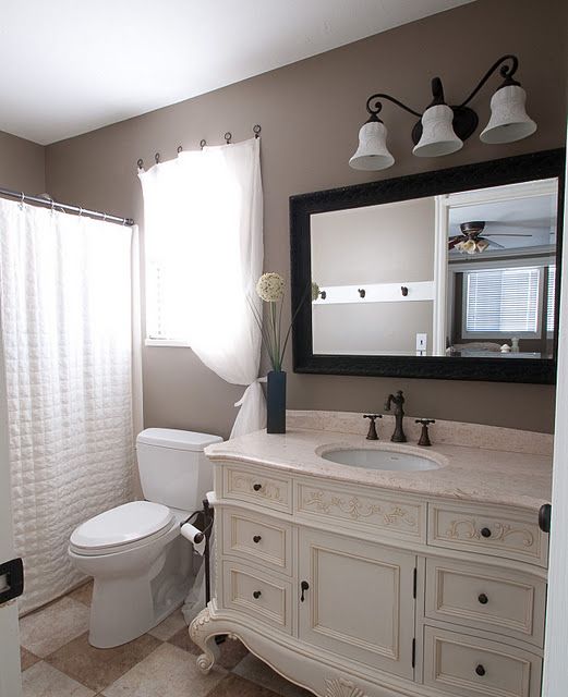 a vintage taupe bathroom with taupe walls, a vintahe ornated vanity, a mirror in a black frame, white appliances and a large sconce