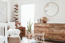 a welcoming and airy modern nursery with a white crub, a leather chair, a stained dresser, a woven pouf, a woven lamp and a stained bookshelf in the corner