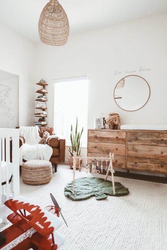a welcoming and airy modern nursery with a white crub, a leather chair, a stained dresser, a woven pouf, a woven lamp and a stained bookshelf in the corner