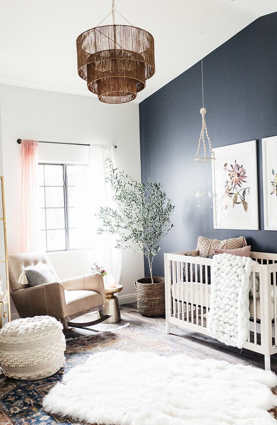 a welcoming boho nursery with a navy accent wall, a white crib and a tan rocker chair, layered rugs, a pendant lamp and some knit pieces