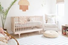 an airy boho nursery with a stained crib, a rattan and upohlstered chair, a fringe decoration, a printed rug plus a potted plant is a cool idea