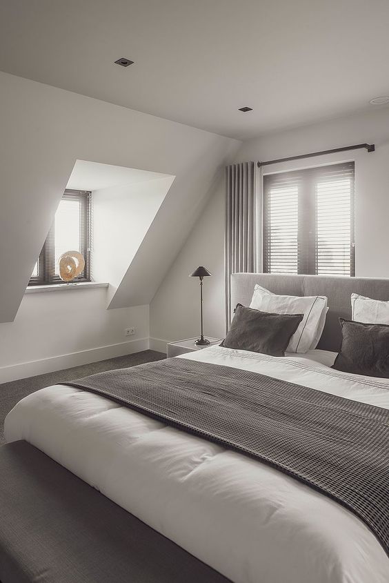 an elegant attic bedroom with windows with dark shades, a grey bed with taupe and white bedding, table lamps
