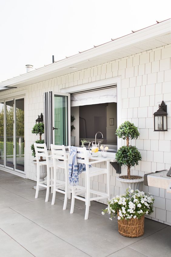a lovely coastal outdoor dining space wiht a pass through window, tall white stools and a floating table, which is a windowsill