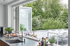 08 a modern kitchen with light-stained cabientry, dark countertops, a white folding window to outdoors that makes bringing food to the outdoor dining space easier