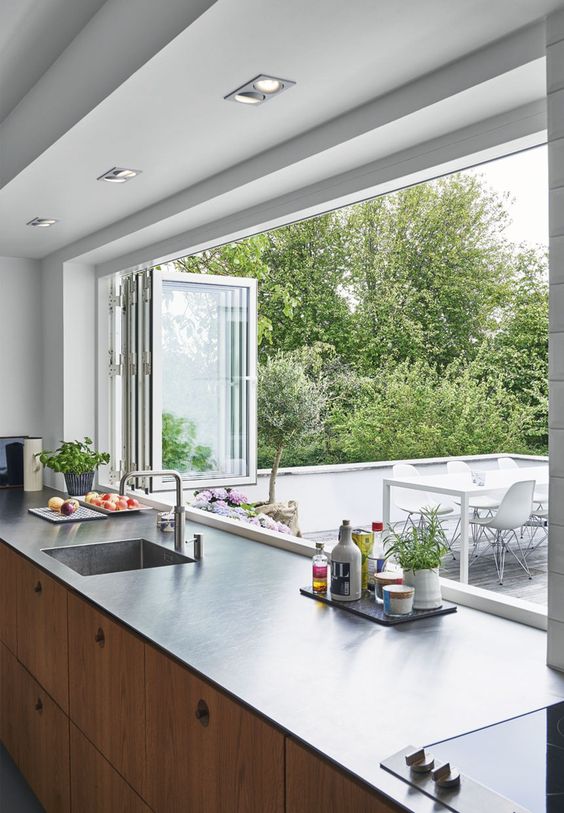 a modern kitchen with light-stained cabientry, dark countertops, a white folding window to outdoors that makes bringing food to the outdoor dining space easier