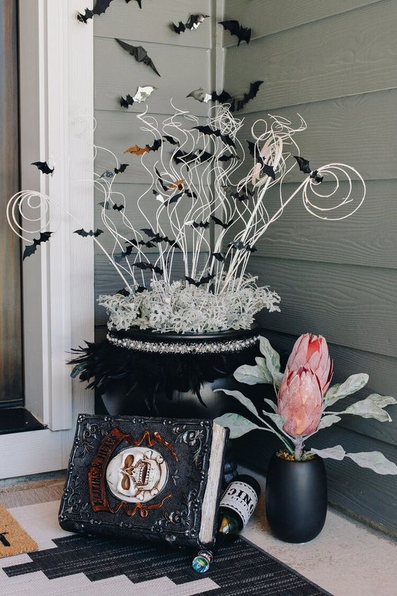 crazy Halloween front porch decor with a spellbook, a black planter with oversized blooms, a black cauldron with twigs and black bats