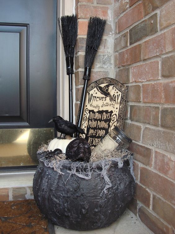 a Halloween porch decoration of an old witch cauldron with a pumpkin, a skull, a jar, a tombstone, a blackbird and some witches' brooms