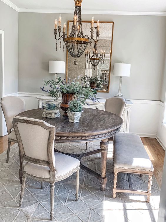 a French country dining room with greige walls, paneling, a fireplace, a stained round table, neutral chairs and a vintage chandelier