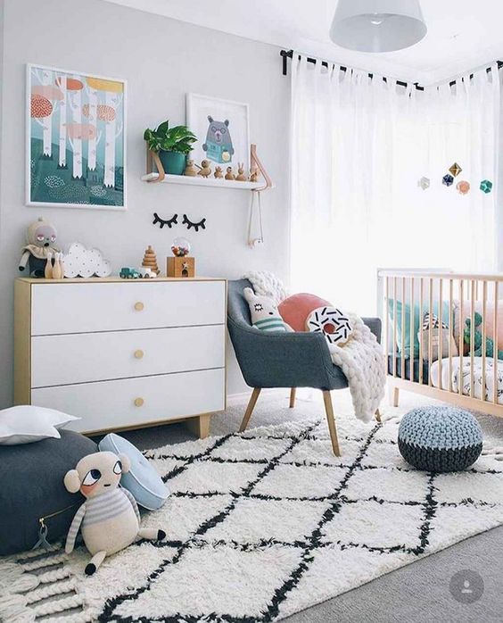 A Scandi nursery with bright touches   artworks, pillows, a pouf and a chair and some bright bedding in the crib is lovely