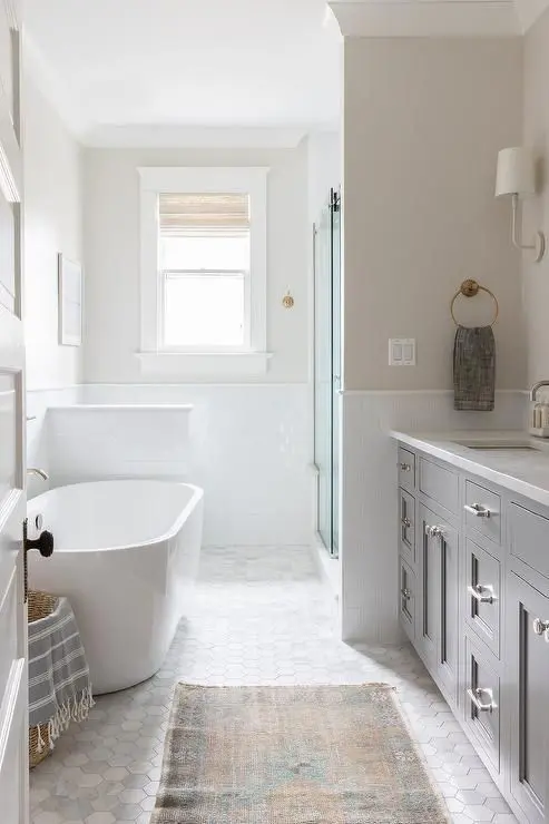 a beautiful farmhouse bathroom with greige walls and a marble hex tile floor, a grey vanity and an oval tub, a shower space