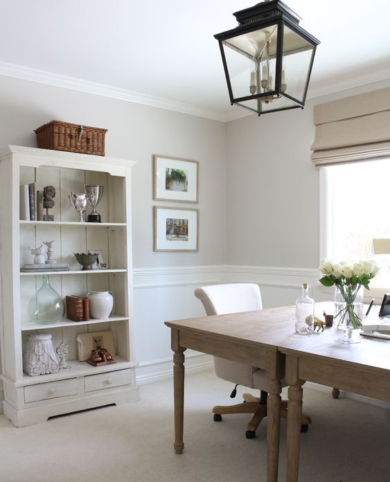 a beautiful farmhouse home office with greige walls and white panels, a neutral planked storage unit, stained tables and a pendant lantern