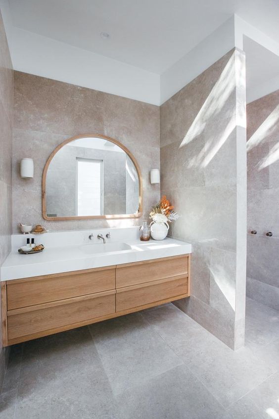 A beautiful greige bathroom clad with large scale tiles, a light stained built in vanity, an arched mirror and a dried leaf arrangement