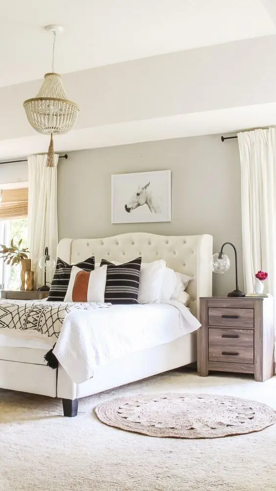 a cozy farmhouse bedroom with greige walls, a creamy upholstered bed, stained nightstands, neutral and printed bedding, a wooden bed chandelier and neutral curtains