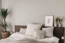 a cozy greige bedroom with a dark upholstered bed, potted plants and blooms, a white nightstand and printed bedding is inviting