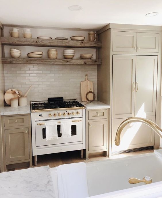 a cozy greige kitchen with shaker cabinets, white stone countertops and a white skinny tile backsplash, reclaimed wood built-in shelves and gold fixtures