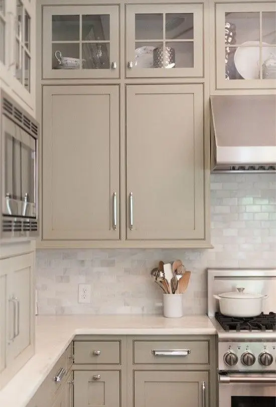 a gorgeous greige kitchen with shaker and glass frame cabinets, a white marble subway tile backsplash and shiny handles