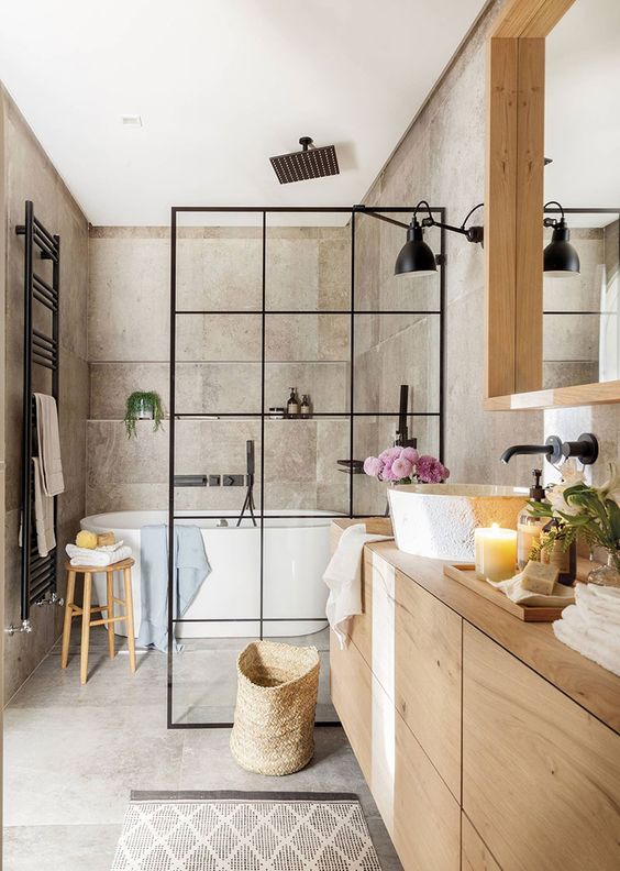 A greige bathroom clad with concrete, with a light stained vanity and a large mirror, a shower space with a black space divider and black fixtures