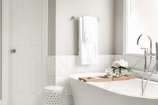 a greige bathroom with white marble tiles, an oval tub by a frosted glass window, a side table and a bathtub caddy is cool