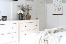 a greige farmhouse bedroom with white paneling, white furniture and textiles, mini artworks and some pretty decor and accessories