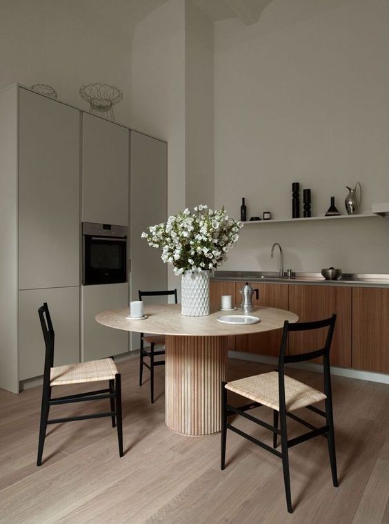 A greige kitchen combined with a kitchen, light stained furniture, a round table and woven chairs, a grey storage unit, a long shelf and some blooms
