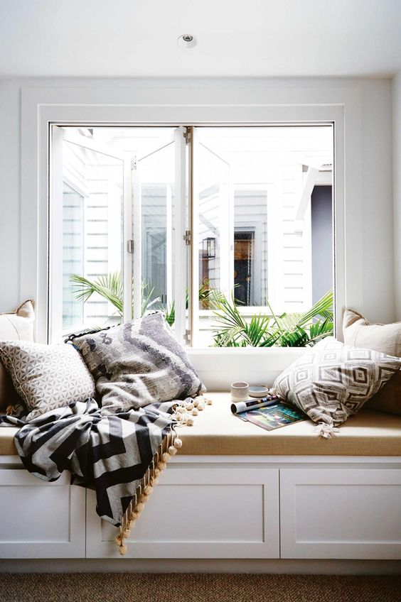 A large window with a built in daybed with a tan cushion and lots of printed pillows and a blanket with pompoms form a cool reading nook